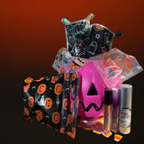A Halloween-themed beauty gift set featuring a pink jack-o'-lantern bucket, black and orange pumpkin-patterned pouches, a spiderweb pouch, a small black ghost bag, and two cosmetic bottles on a dark gradient background.