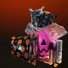 A Halloween-themed beauty gift set featuring a pink jack-o'-lantern bucket, black and orange pumpkin-patterned pouches, a spiderweb pouch, a small black ghost bag, and two cosmetic bottles on a dark gradient background.